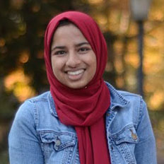 Portrait of Aaruba Ayesha. She is smiling at the camera, wearing a jean jacket and a red headscarf.