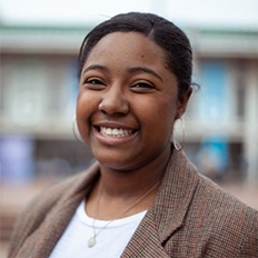 Portrait of Darian Abernathy. Darian is a black woman with straight black hair tied back. She is wearing a white shirt, brown blazer and silver necklace and is smiling at the camera.