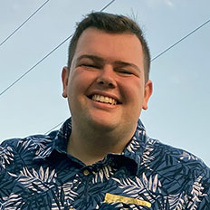 Portrait of Justis Mitchell. Justis is a white man with short brown hair, he is wearing a blue Hawaiian shirt and smiling at the camera.