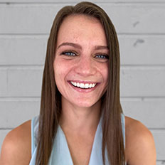 Portrait of Mady Clahane. Mady is a white woman with straight brown hair standing in front of a clapboard wall, wearing a sleeveless blue shirt.