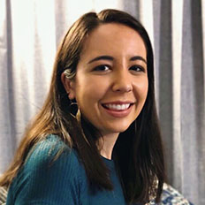 Portrait of Sydney Gutierrez. Sydney is a latina woman with straight brown hair. She is wearing a green shirt and smiling at the camera, sitting in front of a gray curtain.