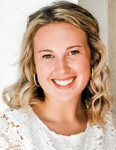 Portrait of Caitlyn Frye. Caitlin is a white woman with wavy white hair. She is wearing a white lace blouse and smiling.