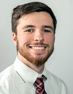 Portrait of Doug Thom. Doug is a white man with brown hair and a brown beard. He is wearing a white button-down shirt, a red plaid tie, and is smiling.