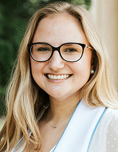 Portrait of Emma Bumgardner. Emma is a white woman with straight blond hair. She is wearing glasses and smiling at the camera.