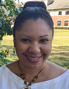 Portrait of Christina Garner. Christina is a black woman with straight black hair tied back with a headband. She is wearing a white shirt and beaded necklace and smiling.