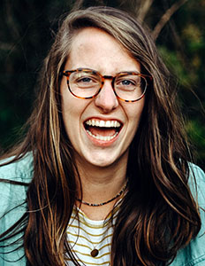 Portrait of Hailey Hawkins. Hailey is a white woman with straight brown hair. She is wearing a striped white shirt, jean jacked and tortoise shell glasses and laughing.