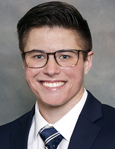 Portrait of JJ Osborn. JJ is a white man with short brown hair. He is wearing a blue suit, blue tie, and brown glasses and smiling.