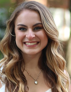 Portrait of Maria Oliva. Maria is a white woman with wavy brown and blond hair. She is wearing a white dress and smiling.