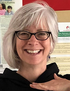 Portrait of Mandy Moore. Mandy is a white woman with gray and white hair, wearing black glasses and a black sweatshirt and smiling.