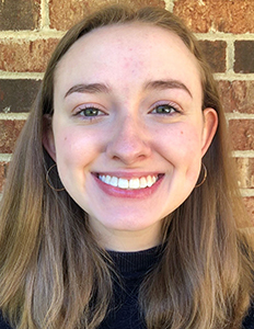 Portrait of Riley Schoonover. Riley is a white woman with straight brown hair. She is wearing a black shirt and smiling in front of a brick wall.