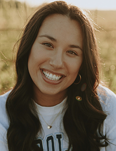 Portrait of Sydney Tanguilig. Sydney is a medium skinned woman with wavy brown hair. She is wearing a white carolina sweatshirt and sitting in a field.