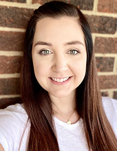 Portrait of Ashley Moore. Ashley is a white woman with straight brown hair. She is smiling, wearing a white shirt and standing in front of a brick wall.