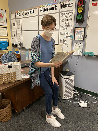 Samantha Shaw, sitting on edge of desk, reading