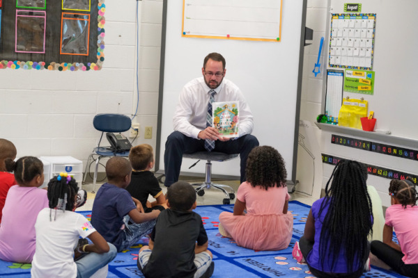 Daniel Watson, principal of Carolina Community Academy, sits in a classroom reading a picture book titled Our School Is a Family to a group of young students seated on a colorful rug during the first day of school in August 2024.