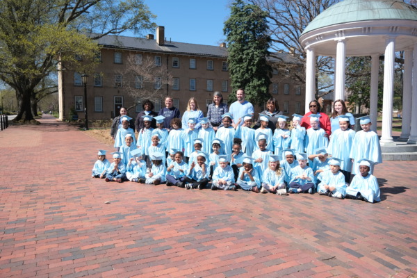 A large group of Carolina Community Academy kindergarten graduates wearing Carolina blue caps and gowns pose for a photo at the Old Well on the UNC-Chapel Hill campus with principal Daniel Watson, UNC School of Education Dean Jill V. Hamm, and CCA staff members during spring 2025.
