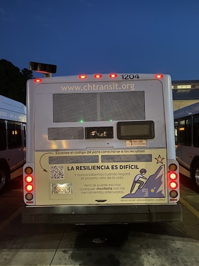 The back of a Chapel Hill Transit bus displays a Spanish-language version of the resiliency campaign ad. The sign reads “La resiliencia es difícil” with an illustration of a person climbing a mountain toward a star. A QR code links to mental health resources, and text along the bottom lists “Apoyo social, Aterrizamiento, Gratitud.”