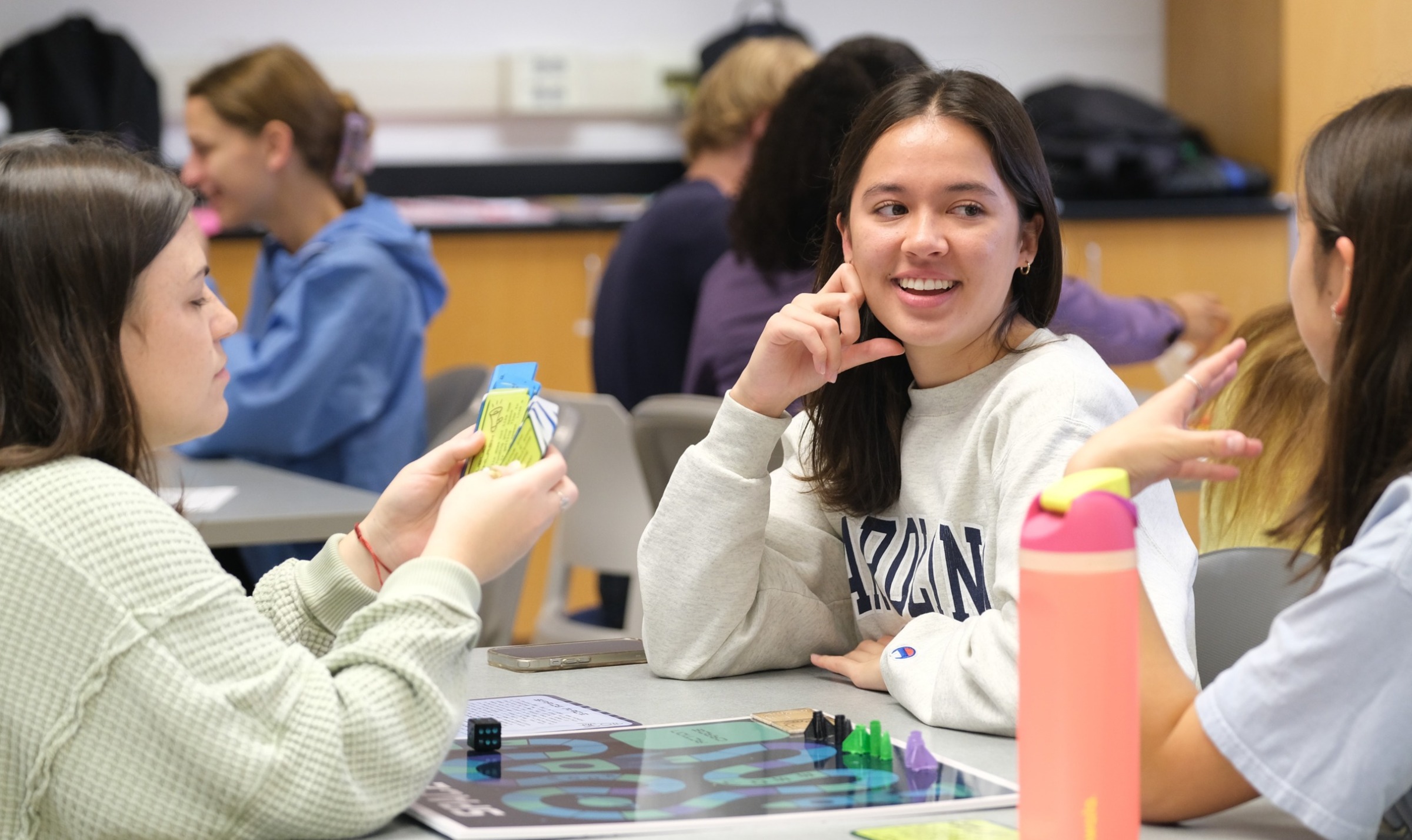 Three students sit at a table playing a board game with cards.