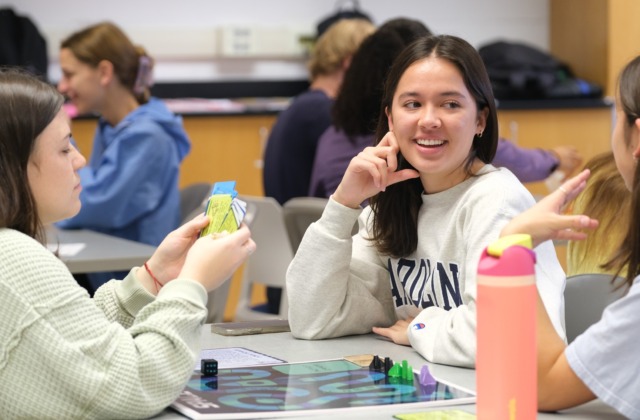 Three students sit at a table playing a board game with cards.