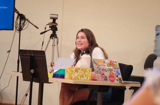 School counseling student Chloe Rhodes sits at a table with her laptop open, presenting to the cast of The Wolves about neuroscience