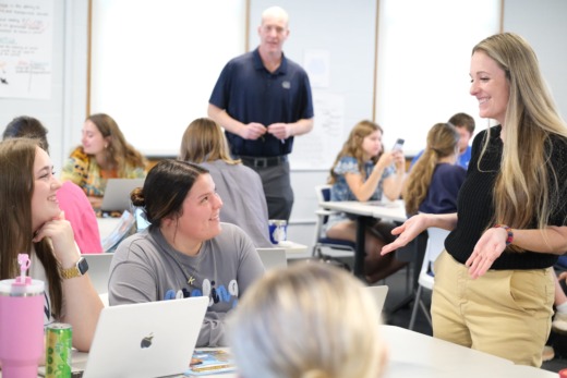 Nicole Danilo works with two Teaching Fellows during a MAT class. They speak at a table with computers surrounded by other students working.