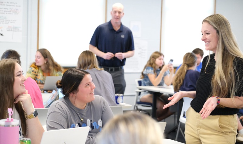 Nicole Danilo works with two Teaching Fellows during a MAT class. They speak at a table with computers surrounded by other students working.