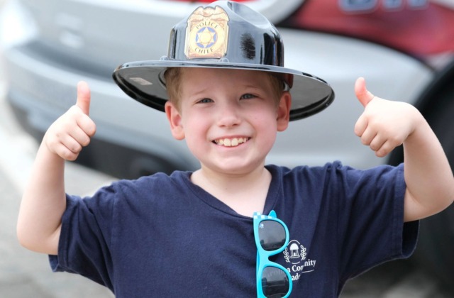 Smiling Carolina Community Academy student wearing a black police chief helmet gives two thumbs up while participating in a community partner activity during CCA’s inaugural summer camp.