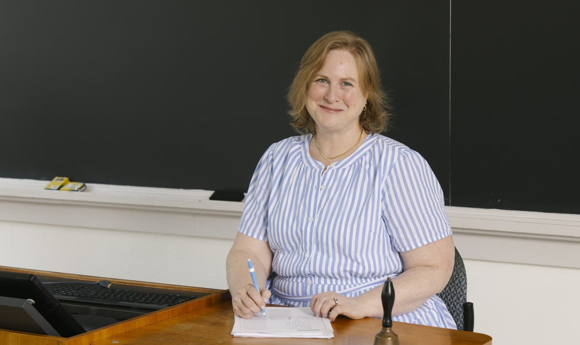 UNC School of Education faculty member Kristin Papoi sits at the front of a classroom, holding a pen beside a stack of papers, with a brass school bell on the desk in front of her.