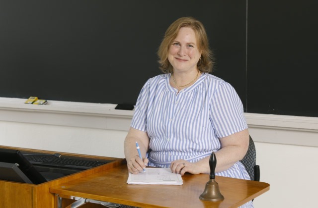 UNC School of Education faculty member Kristin Papoi sits at the front of a classroom, holding a pen beside a stack of papers, with a brass school bell on the desk in front of her.