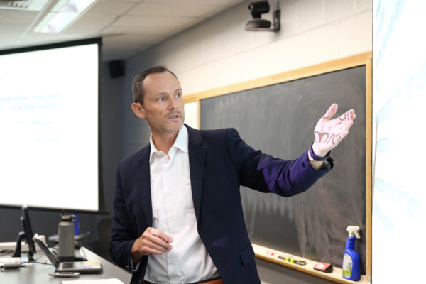 Todd Jensen, Ph.D., stands at the front of a classroom, gesturing toward a projected presentation while teaching. A chalkboard and presentation screen are visible behind him.