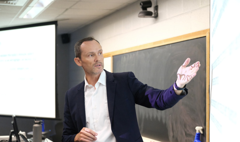 Todd Jensen, Ph.D., stands at the front of a classroom, gesturing toward a projected presentation while teaching. A chalkboard and presentation screen are visible behind him.